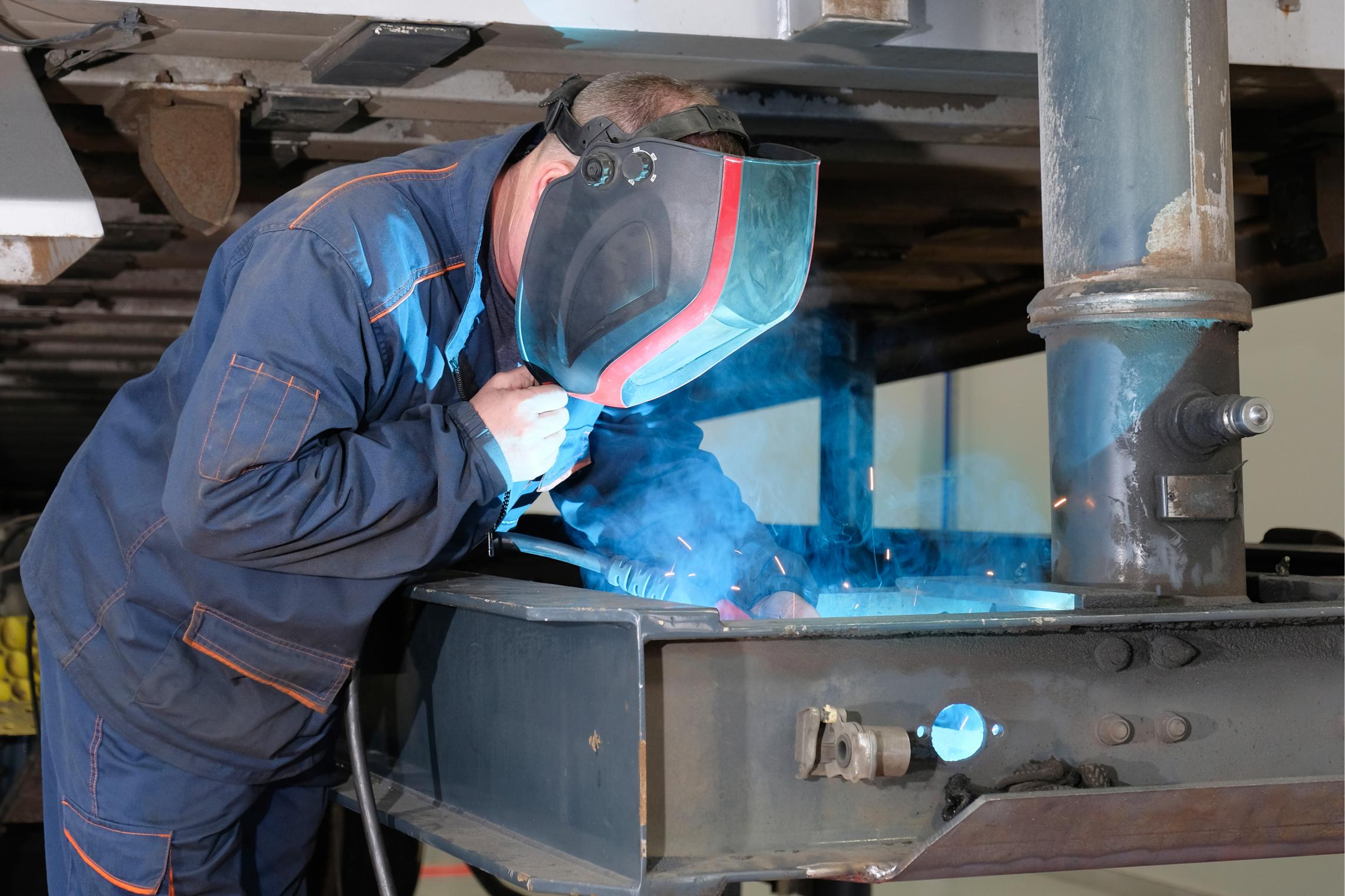 A welder in a helmet works in a garage, welding a cargo trailer. Service and repair of trucks - Copy